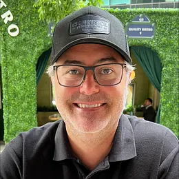 Smiling man wearing glasses and a black cap in front of greenery and a restaurant entrance