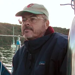 man wearing a cap and jacket sitting on a boat near calm water with trees in the background