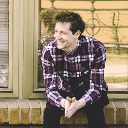 Smiling person wearing a plaid shirt sitting by a window with hands clasped on their knees