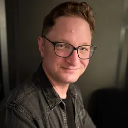 A close-up portrait of Jay Gilmore, a man with short brown hair and glasses, wearing a dark denim button-down shirt over a black t-shirt, smiling warmly against a dark background.